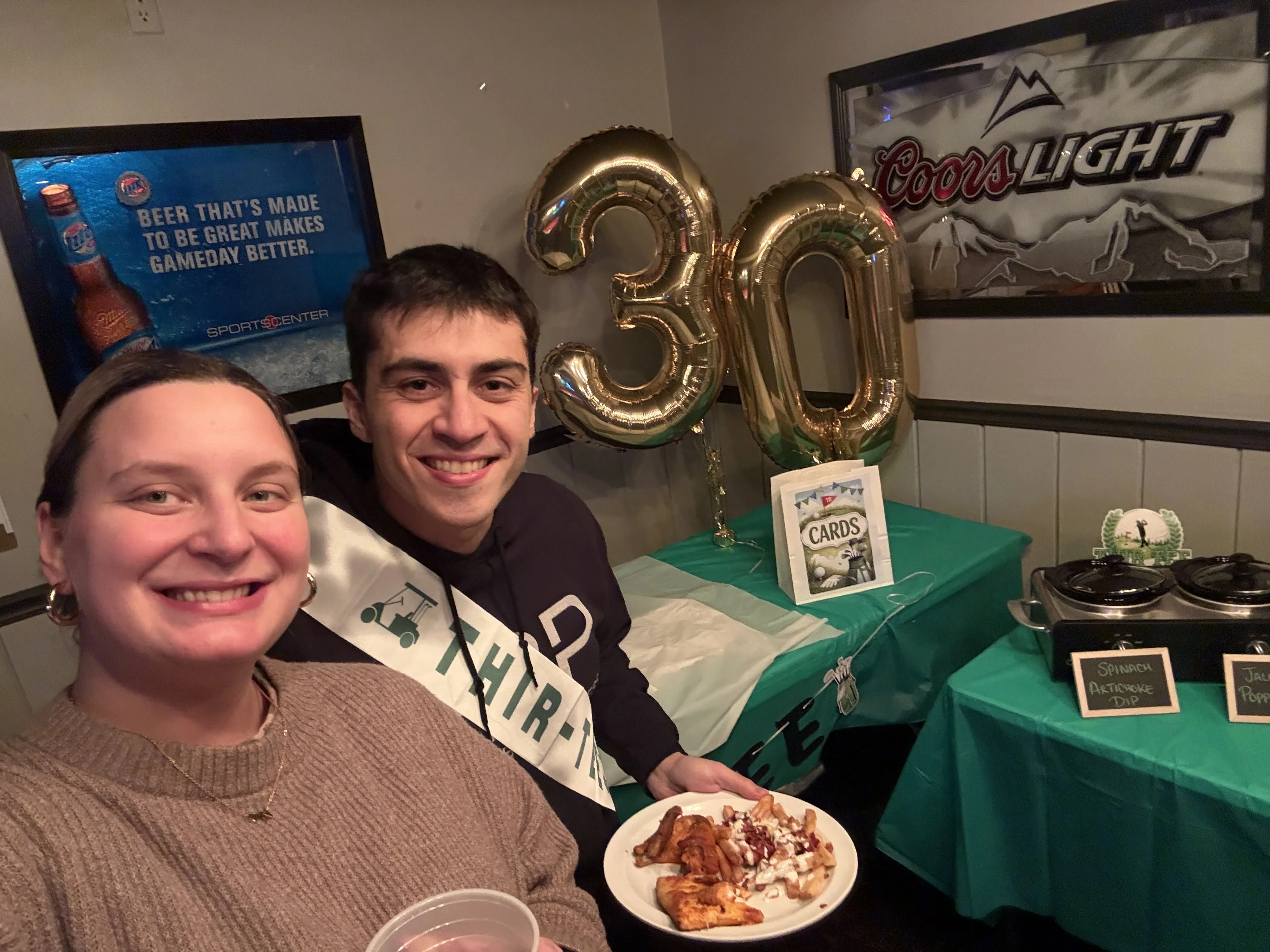 Amanda and Mark at his surprise 30th birthday party, Mark in a THIR-TEE golf sash beside gold 3 and 0 balloons.