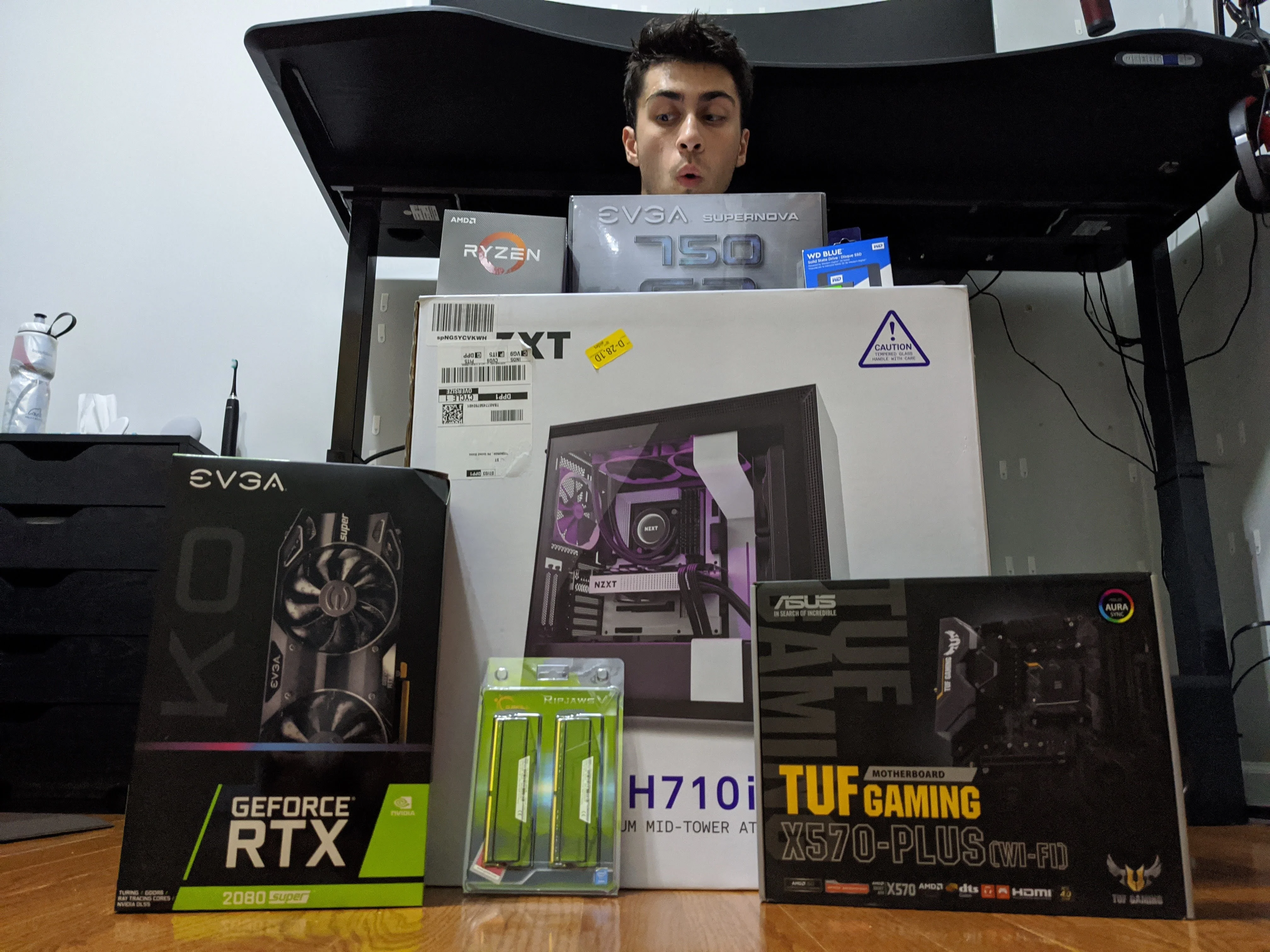 Mark peeking over a stack of boxed computer parts, including an NZXT case, an RTX 2080 Super, and a Ryzen processor.