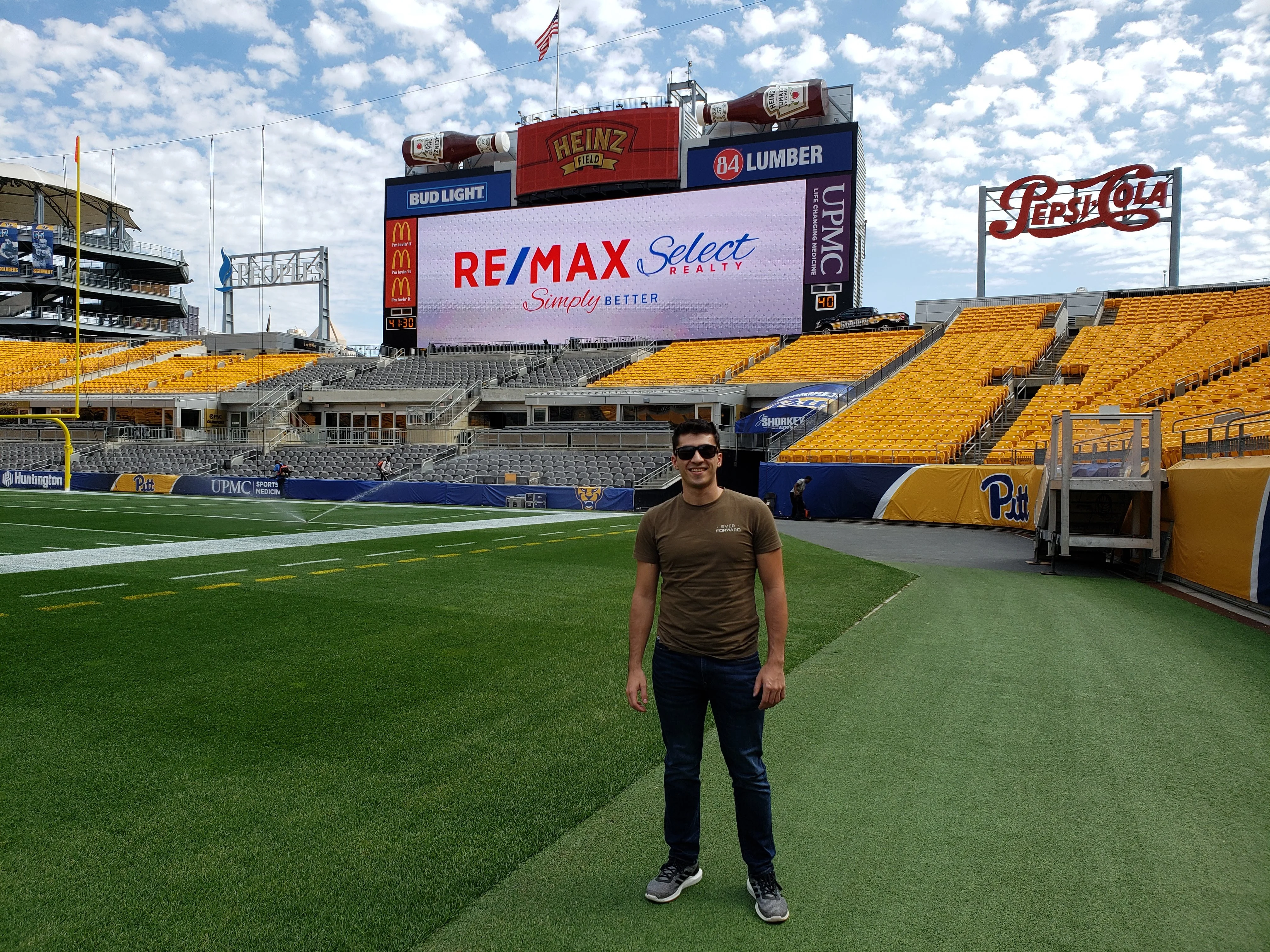 Mark standing on the field at Heinz Field with the RE/MAX Select Realty logo on the stadium scoreboard behind him.