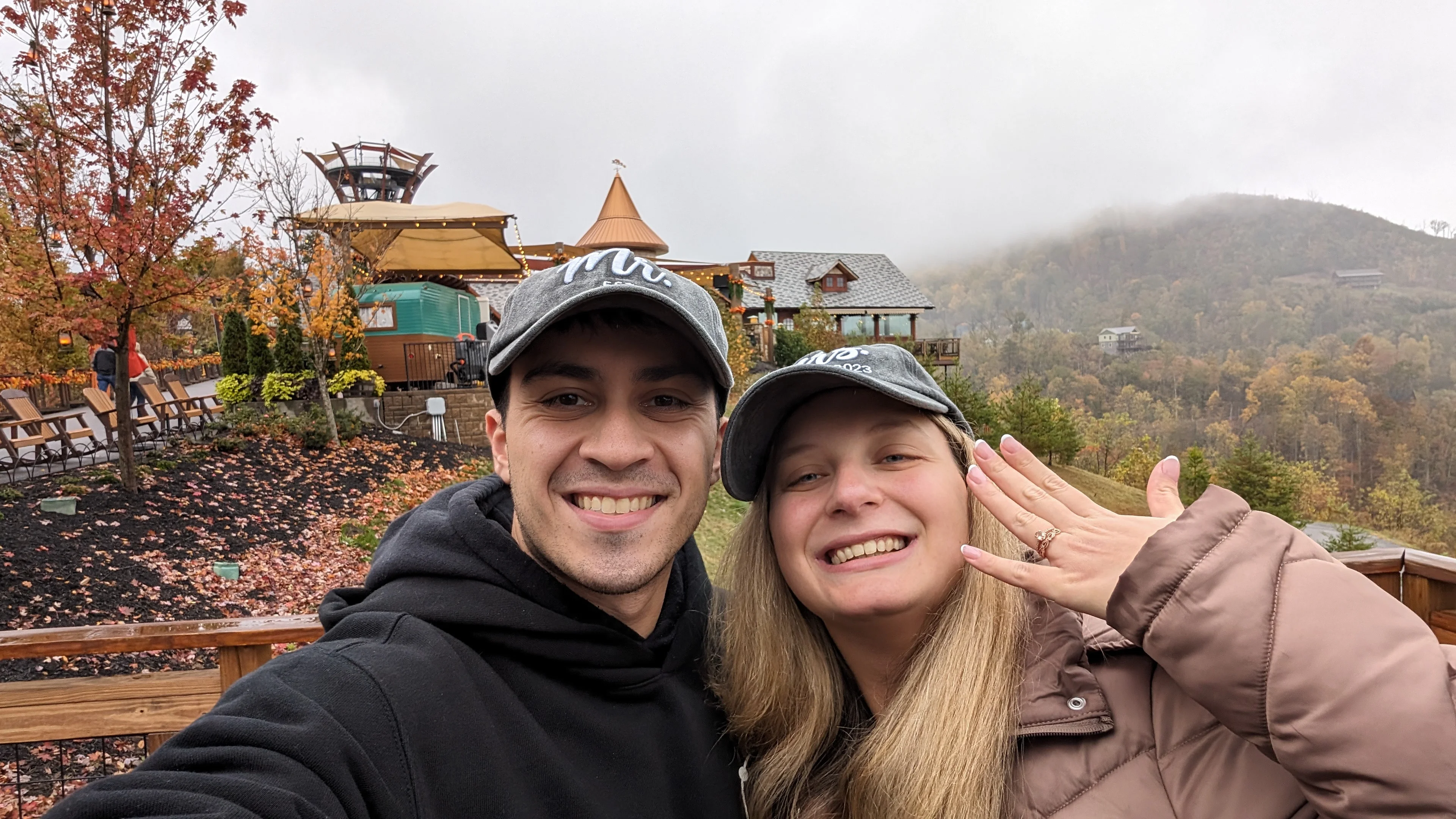 Mark and Amanda in matching Mr. and Mrs. hats taking a selfie on a foggy fall mountaintop, Amanda showing her ring.