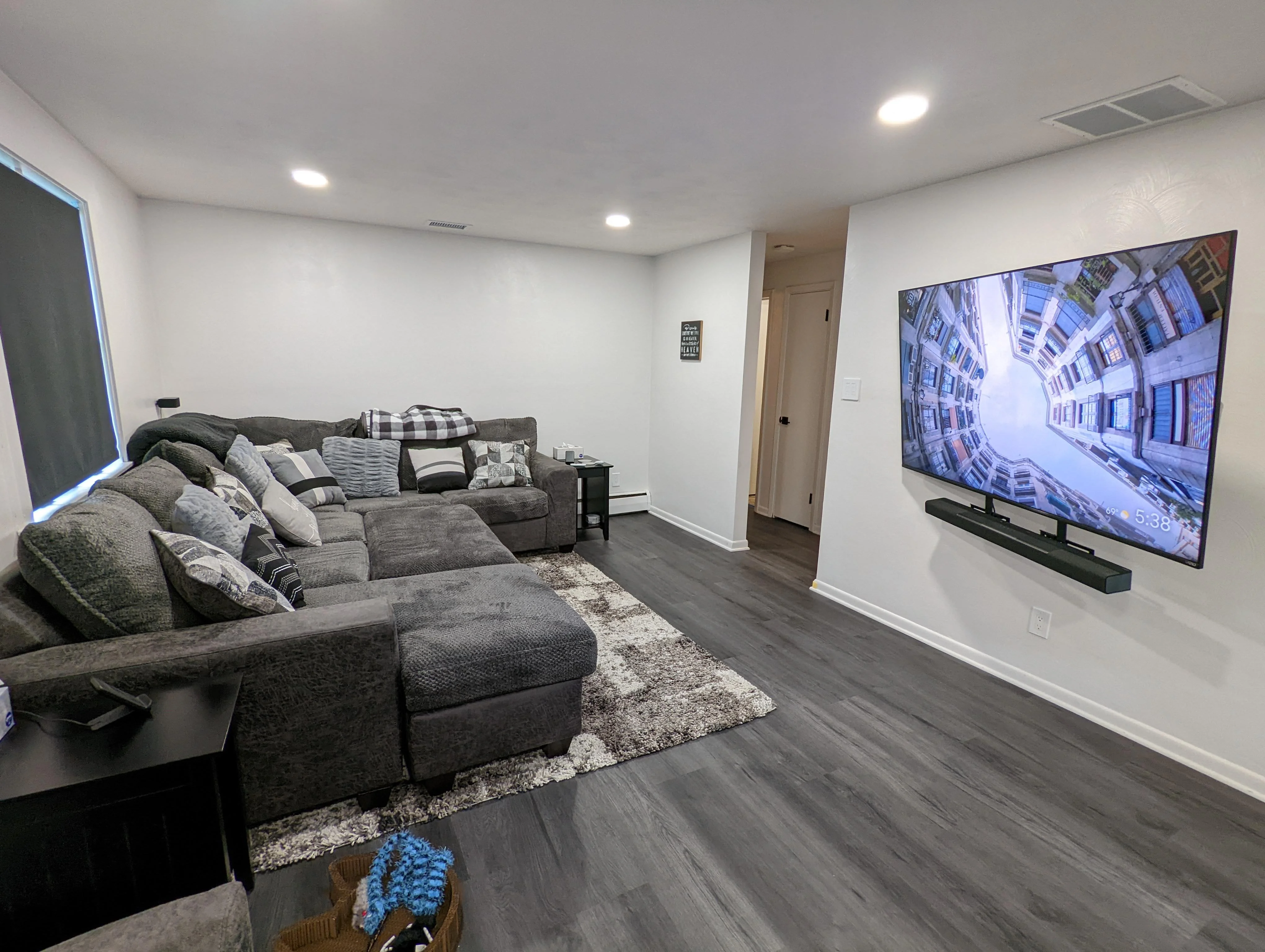 The living room after the renovation: white walls, gray vinyl plank floor, recessed lights, a sectional, and a wall-mounted TV.