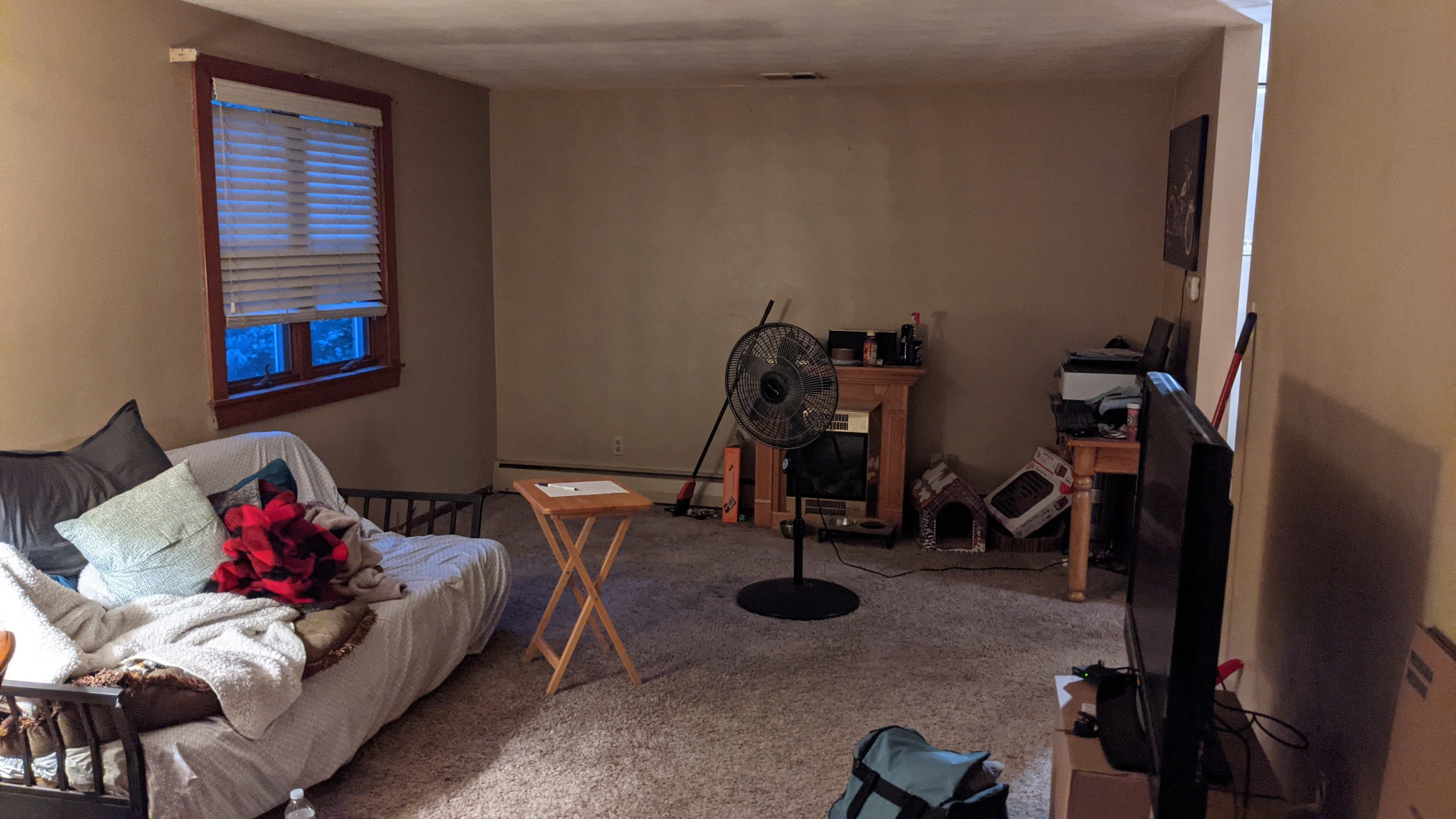 The living room before the renovation: tan walls, beige carpet, a futon, and a cluttered corner with a fan and folding table.