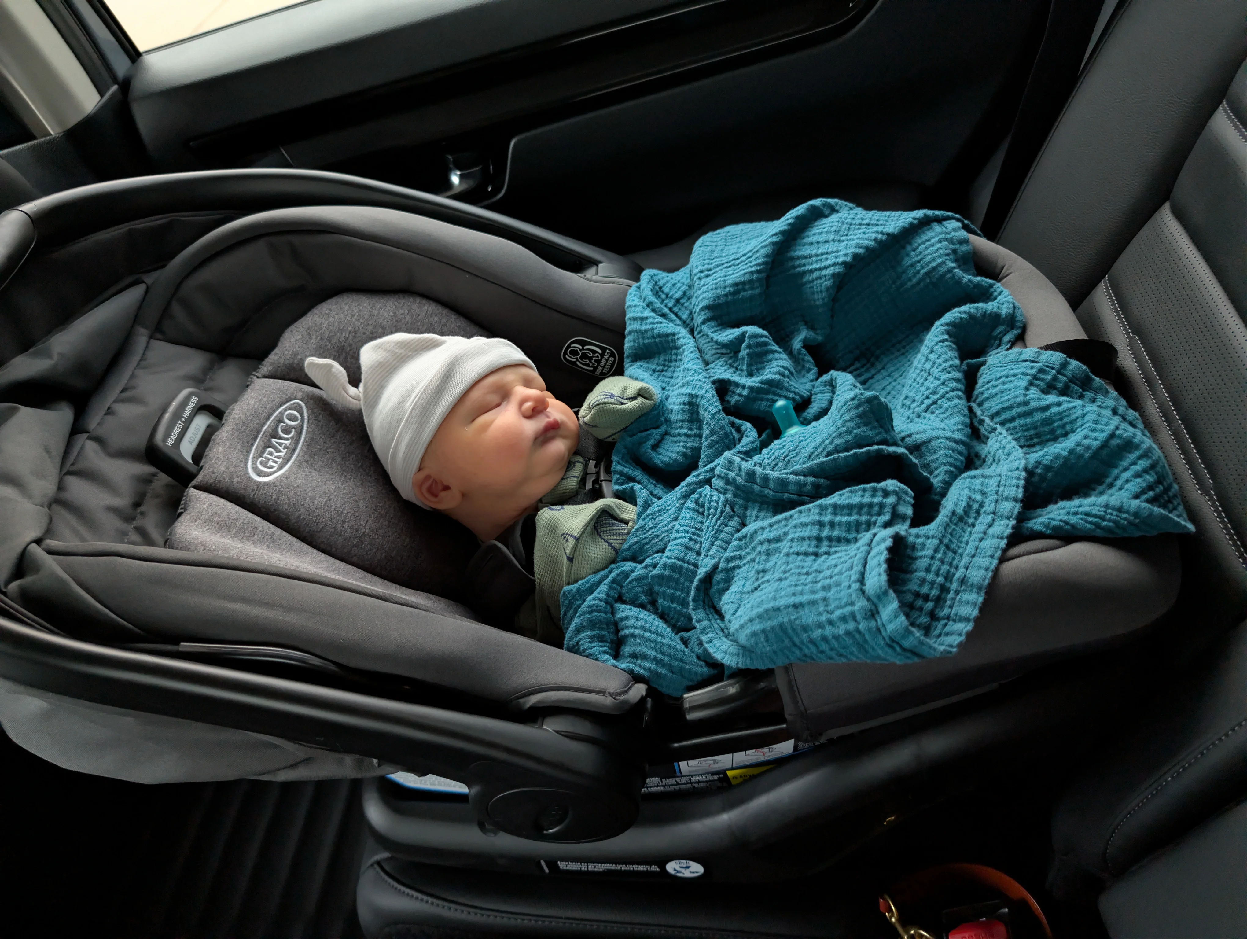 Nolan asleep in a car seat in a knit hat with a teal blanket, on the ride home from the hospital.