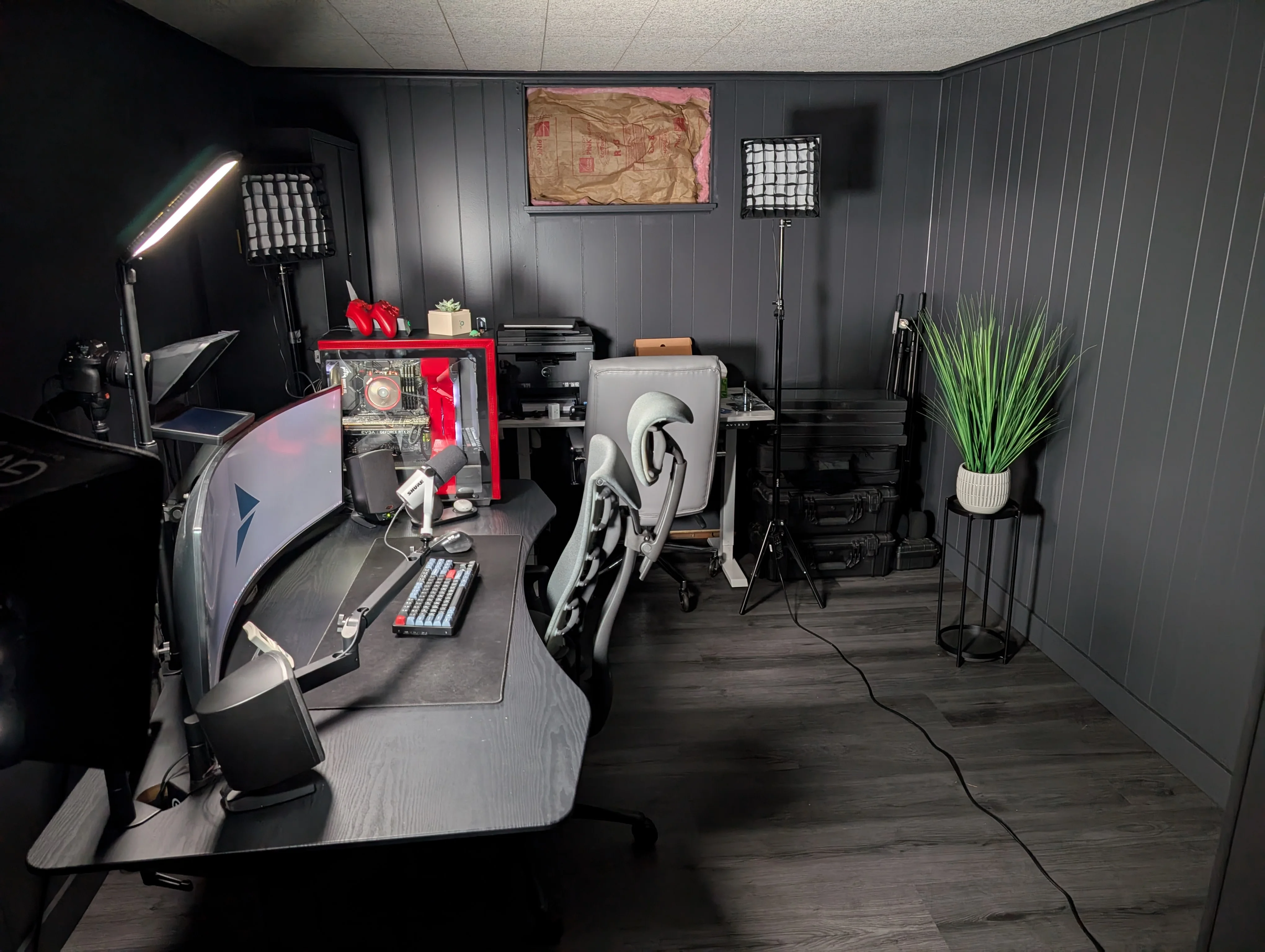 The finished office and studio: dark painted walls, gray vinyl plank flooring, a curved desk setup, studio lights, and a red PC.