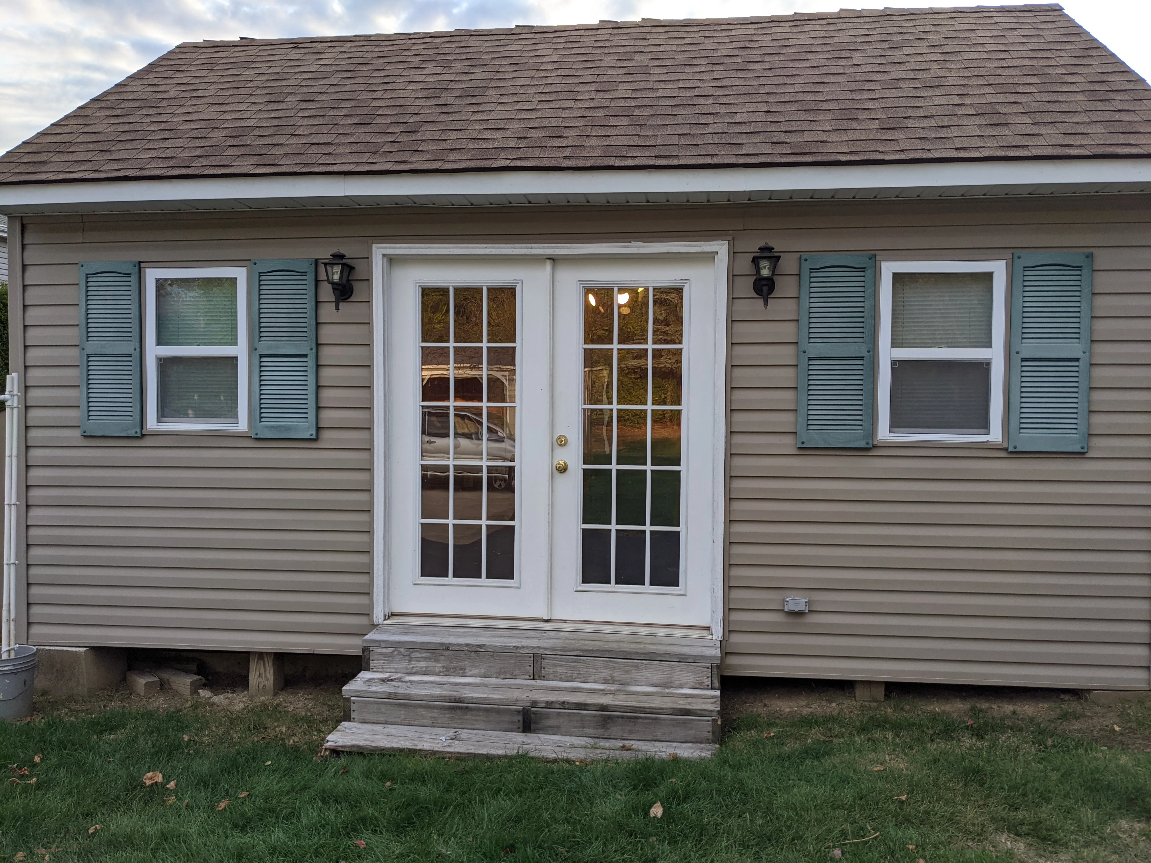 A tan shed with French doors, green shutters, and wooden steps, lit from inside at dusk.