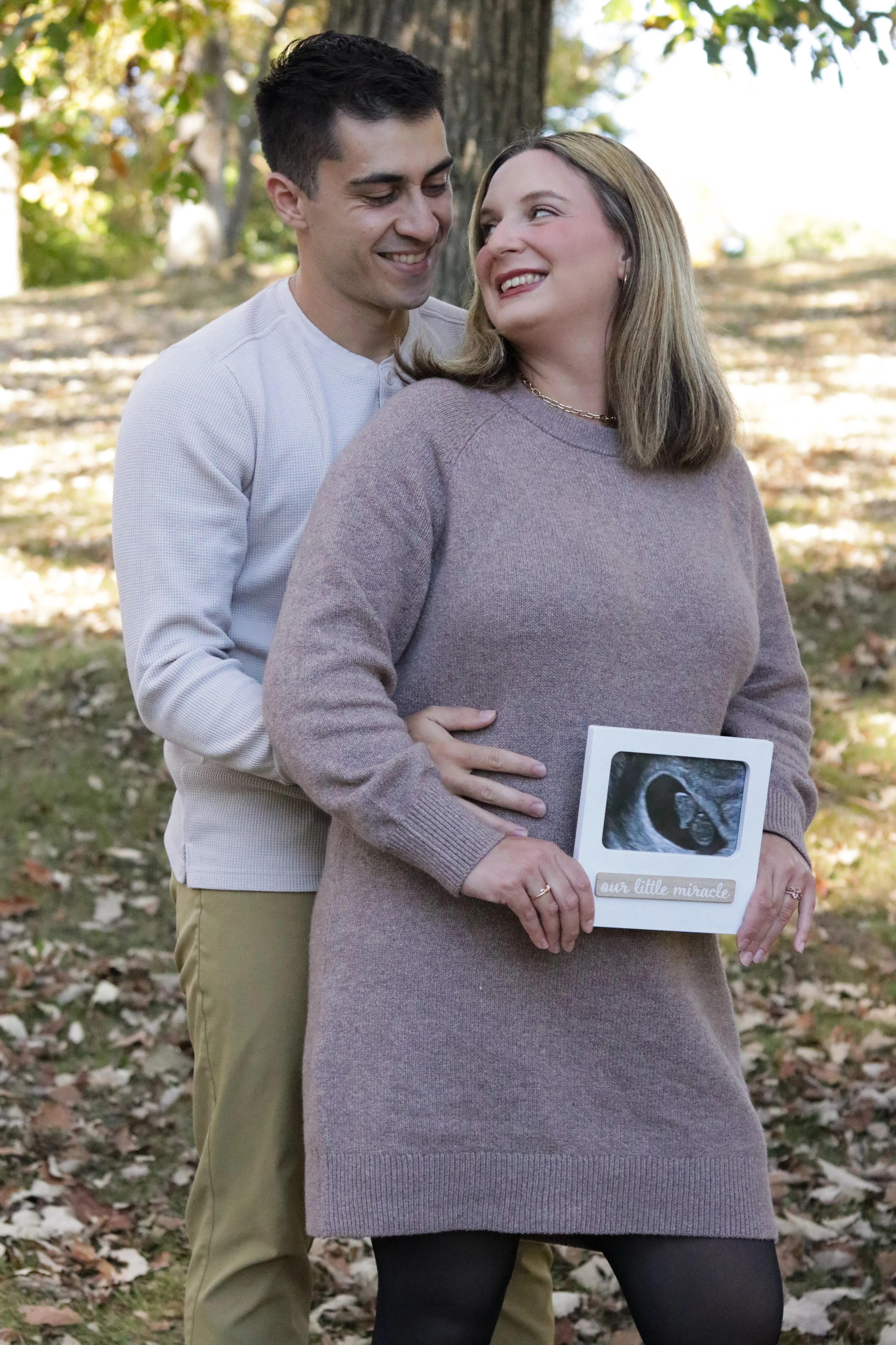 Mark and Amanda outside among fall leaves, Amanda holding a framed ultrasound that reads our little miracle.