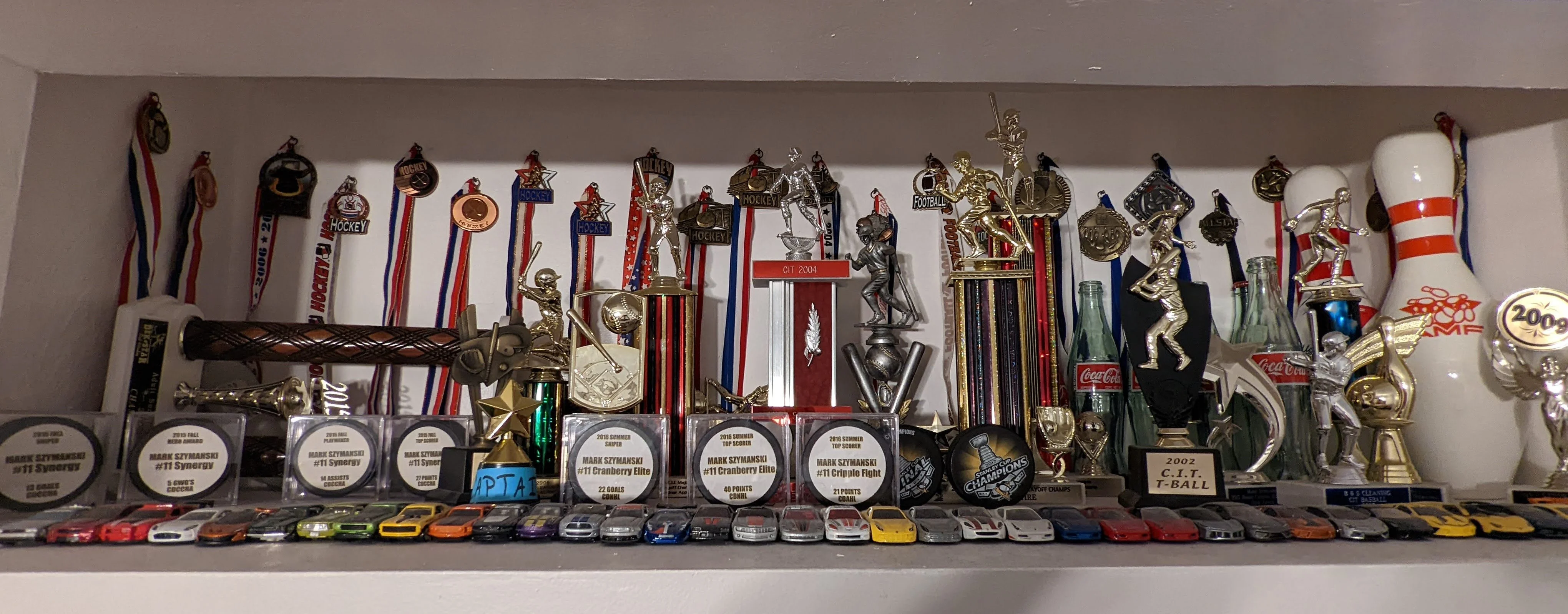 My full trophy shelf: hockey and football medals, C.I.T. baseball trophies, bowling trophies and an AMF pin, engraved hockey pucks, and a row of toy cars in front.